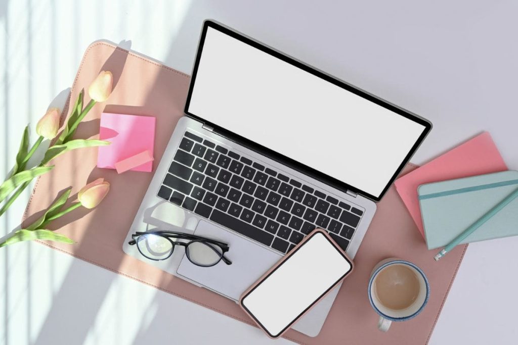 A laptop and smartphone sitting on a desk with coffee, glasses, notebooks, and flowers.