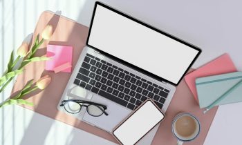 A laptop and smartphone sitting on a desk with coffee, glasses, notebooks, and flowers.