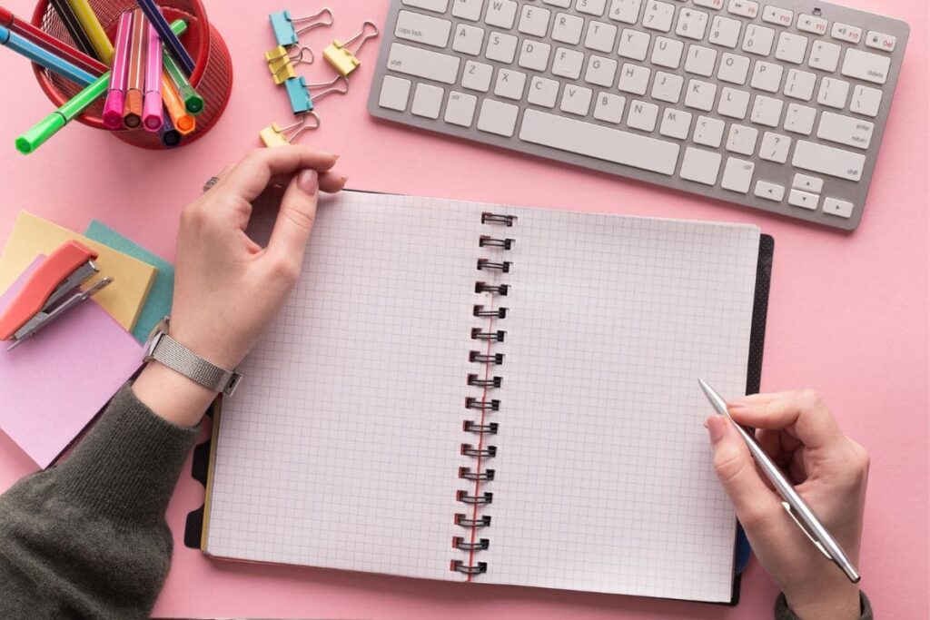 Overhead view of a woman's hands writing in a notebook while sitting at a desk.