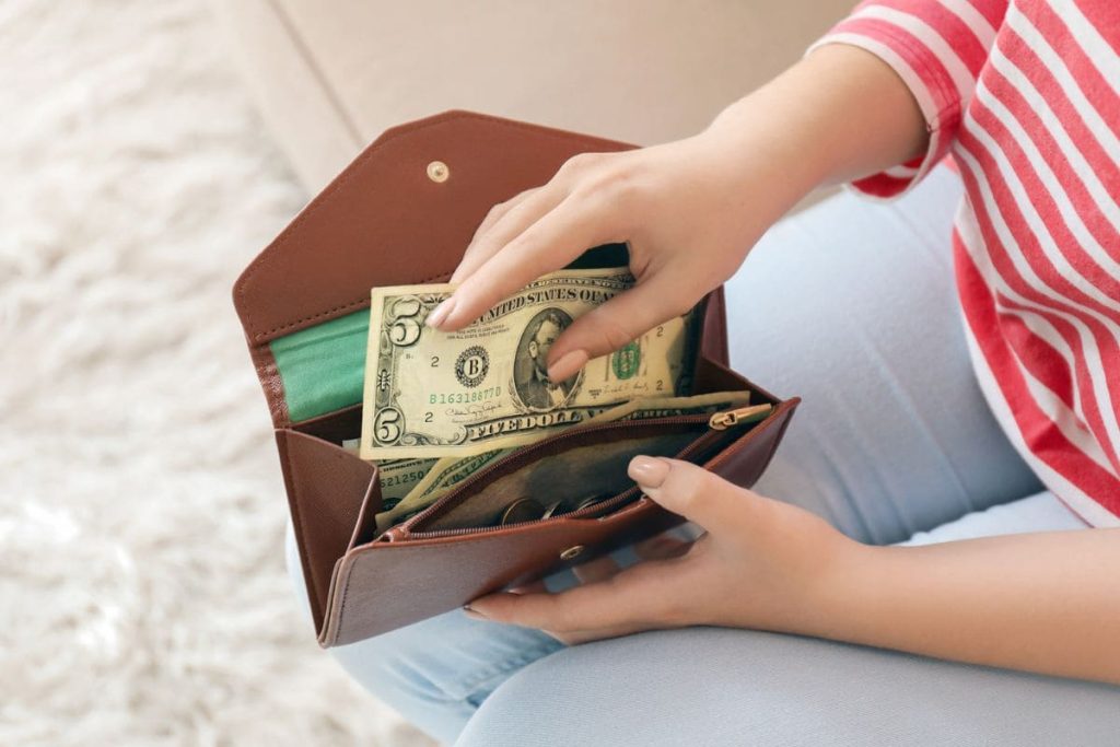 A woman, holding a wallet and putting a $5 bill into it.