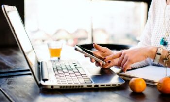 A woman holding a smart phone while working on a laptop at home.