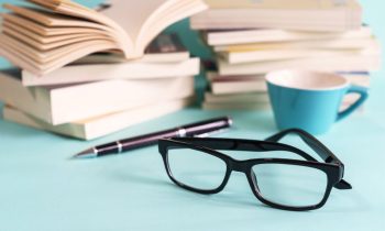 A stack of books on a desk next to a pair of glasses and a cup of coffee.