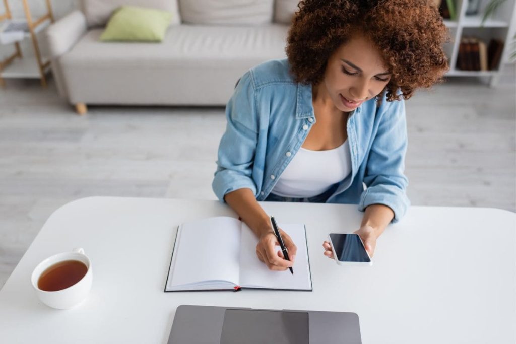 A woman writing in a notebook and working on her LinkedIn profile at a home office desk.