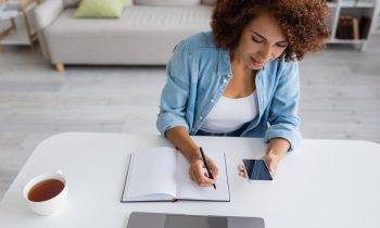 A woman writing in a notebook and working on her LinkedIn profile at a home office desk.