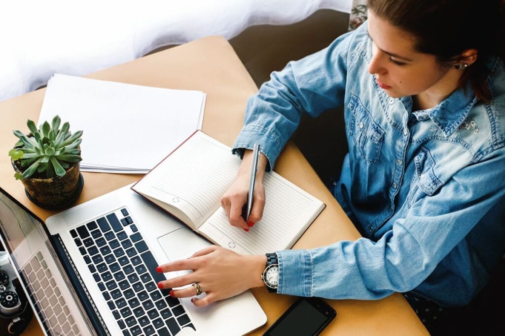 Overhead view of a woman sitting at a home office desk, using a laptop, and writing in a notebook.