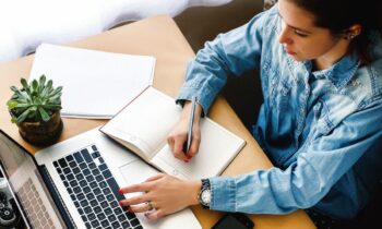 Overhead view of a woman sitting at a home office desk, using a laptop, and writing in a notebook.