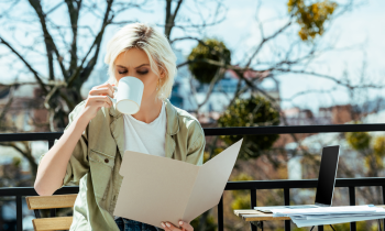 woman with blonde hair outdoors drinking coffee and reading a file folder with documents