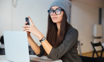 Young woman wearing glasses and a hat holding her smartphone looking at her laptop for ways to make money online