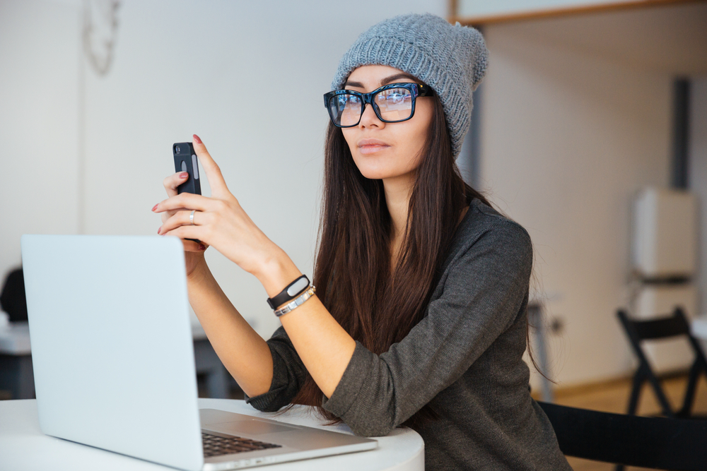 Young woman wearing glasses and a hat holding her smartphone looking at her laptop for ways to make money online